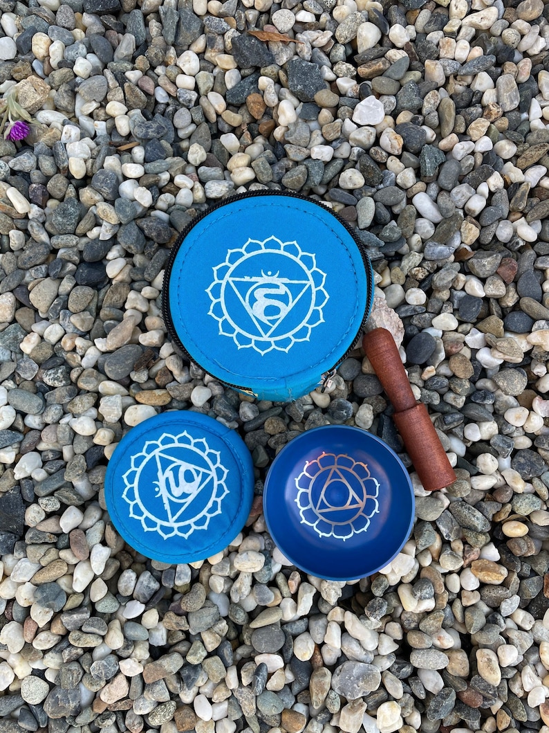 May include: Three blue singing bowls with a white design of a chakra symbol. The bowls are on a bed of small gray and white stones. A wooden striker is included.