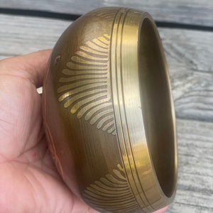 Puede incluir: Un cuenco cantor de color dorado con intrincados dise&ntilde;os grabados. El cuenco es redondo y tiene un borde ligeramente acampanado.