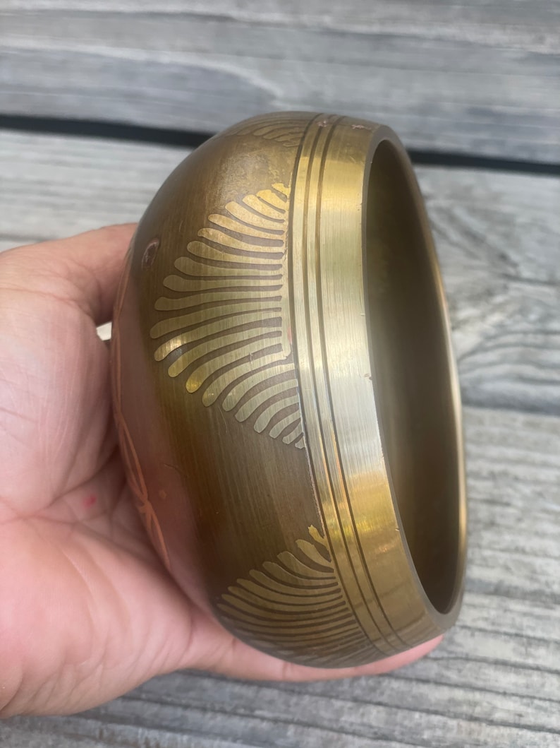 Puede incluir: Un cuenco cantor de color dorado con intrincados dise&ntilde;os grabados. El cuenco es redondo y tiene un borde ligeramente acampanado.