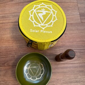 May include: A yellow fabric pouch with a zipper closure and the text "Solar Plexus" printed on it. The pouch contains a green singing bowl with a white symbol and a wooden striker.