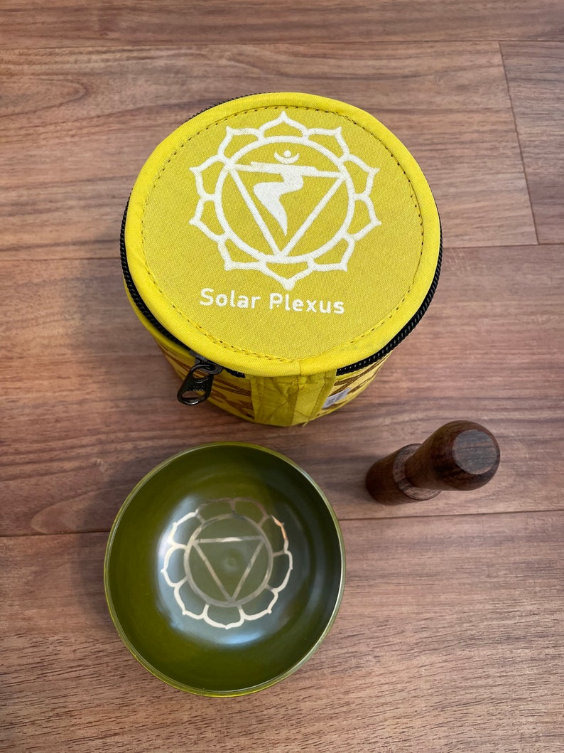 May include: A yellow fabric pouch with a zipper closure and the text "Solar Plexus" printed on it. The pouch contains a green singing bowl with a white symbol and a wooden striker.
