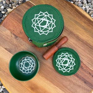 May include: A green singing bowl set with a wooden striker, a green fabric carrying case, and a green felt cushion. The bowl, case, and cushion all feature a white symbol of the heart chakra.