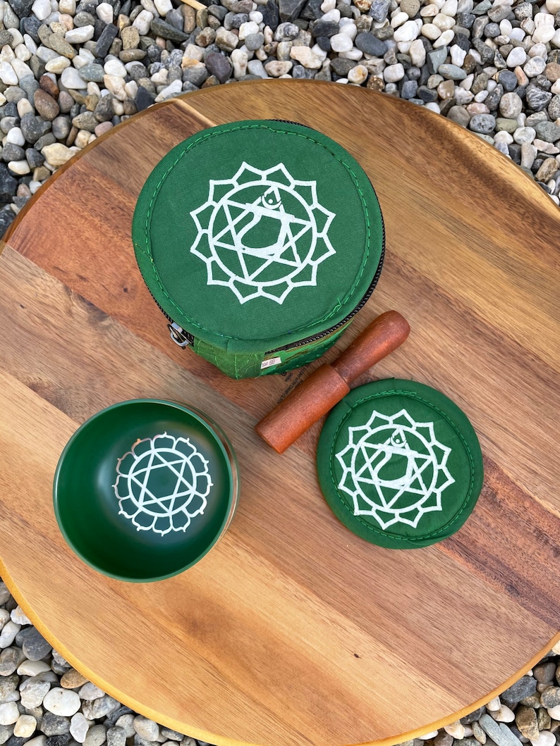 May include: A green singing bowl set with a wooden striker, a green fabric carrying case, and a green felt cushion. The bowl, case, and cushion all feature a white symbol of the heart chakra.