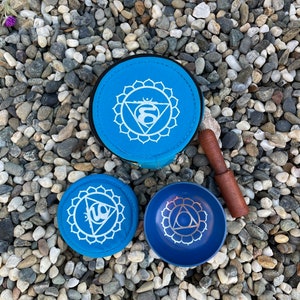 Puede incluir: Tres cuencos cantores azules con un s&iacute;mbolo blanco en cada uno. Los cuencos est&aacute;n sobre un lecho de peque&ntilde;as piedras grises y blancas. Un cuenco est&aacute; en una bolsa azul con cremallera. Un mazo de madera est&aacute; al lado de los cuencos.