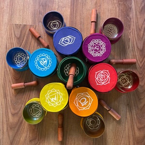 May include: Seven singing bowls with mallets, each with a different color and chakra symbol, arranged in a circle on a wooden surface.