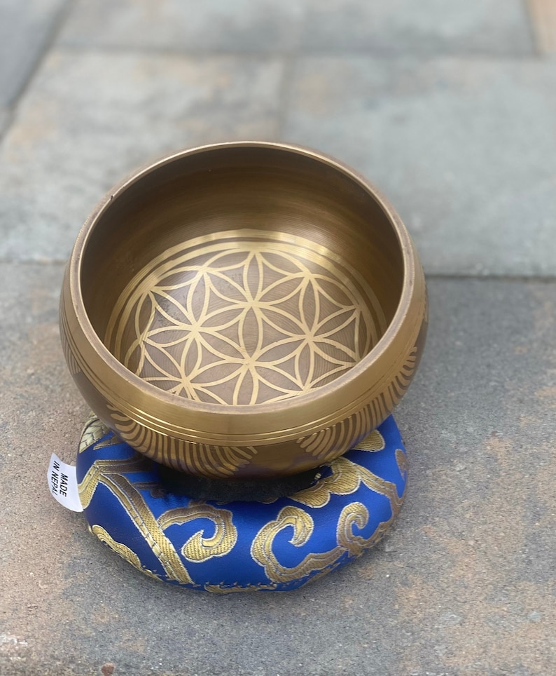 Puede incluir: Un cuenco cantor dorado con un dise&ntilde;o de flor de la vida en su interior. El cuenco est&aacute; sentado sobre un coj&iacute;n azul y dorado con el texto "Hecho en Nepal".