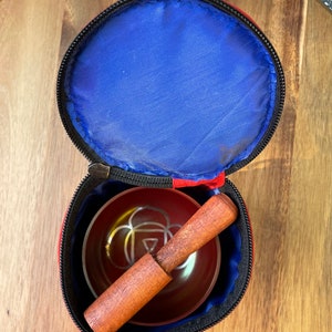 May include: A red and black carrying case with a zipper closure, containing a red singing bowl with a wooden striker. The bowl has a silver design on the inside.