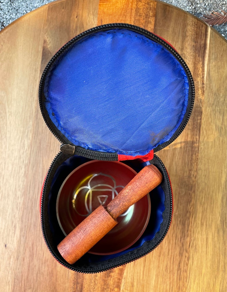 May include: A red and black carrying case with a zipper closure, containing a red singing bowl with a wooden striker. The bowl has a silver design on the inside.