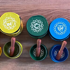Puede incluir: Siete cuencos cantores con mazos de madera, cada uno con un color y un s&iacute;mbolo diferentes que representan los siete chakras. Los cuencos est&aacute;n en fila, de izquierda a derecha: rojo, naranja, amarillo, verde, azul, &iacute;ndigo y violeta.
