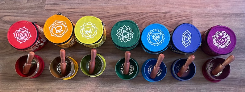 Puede incluir: Siete cuencos cantores con mazos de madera, cada uno con un color y un s&iacute;mbolo diferentes que representan los siete chakras. Los cuencos est&aacute;n en fila, de izquierda a derecha: rojo, naranja, amarillo, verde, azul, &iacute;ndigo y violeta.