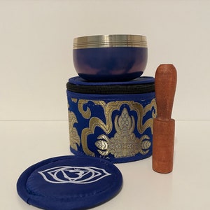 May include: A blue singing bowl with a silver rim and a wooden striker, in a blue and gold patterned carrying case with a zipper closure. The case has a matching blue round cushion with a white symbol.