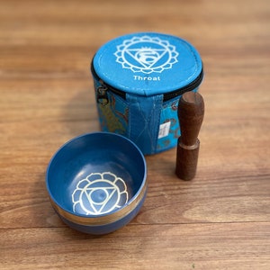 May include: A blue singing bowl with a silver rim and a white symbol of the fifth chakra, the throat chakra, on the inside. The bowl comes with a wooden striker and a blue fabric carrying case with the symbol of the throat chakra and the word "Throat" printed on it.