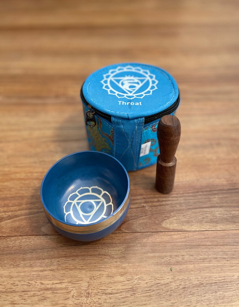 May include: A blue singing bowl with a silver rim and a white symbol of the fifth chakra, the throat chakra, on the inside. The bowl comes with a wooden striker and a blue fabric carrying case with the symbol of the throat chakra and the word "Throat" printed on it.