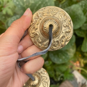 May include: Two small, round, brass cymbals with intricate designs, connected by a black leather strap. The cymbals have a raised centre and are decorated with floral and dragon motifs.