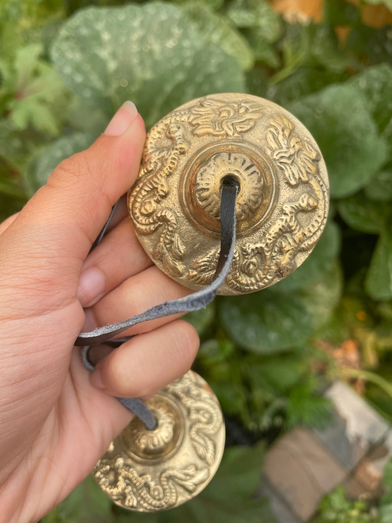 May include: Two small, round, brass cymbals with intricate designs, connected by a black leather strap. The cymbals have a raised centre and are decorated with floral and dragon motifs.