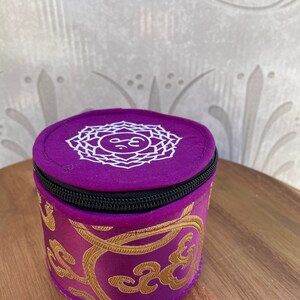 May include: A small purple cylindrical pouch with a zipper closure. The pouch has a white embroidered design of a mandala with the symbol "Om" in the center. The pouch is decorated with gold embroidery.