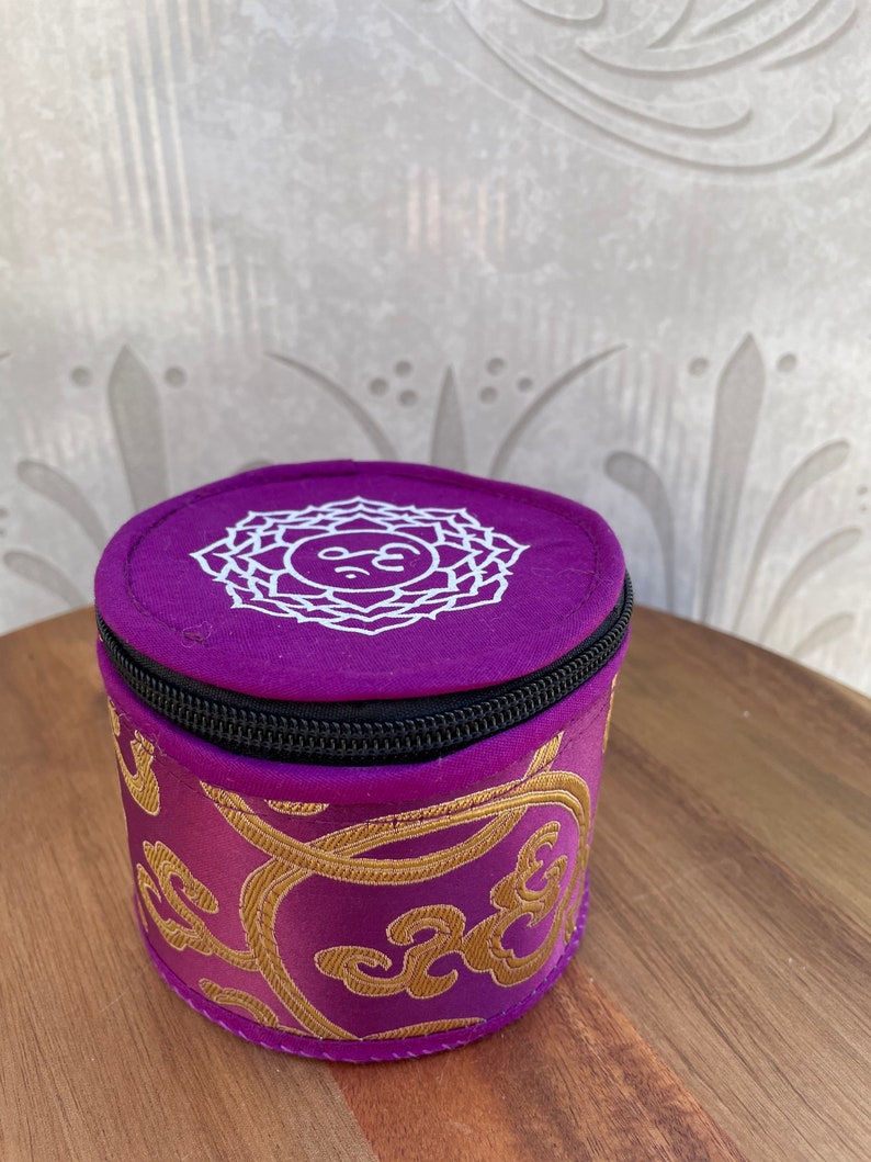 May include: A small purple cylindrical pouch with a zipper closure. The pouch has a white embroidered design of a mandala with the symbol "Om" in the center. The pouch is decorated with gold embroidery.