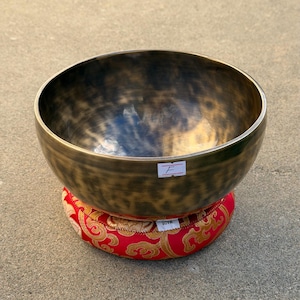 May include: A bronze-colored singing bowl with a dark interior sits atop a red and gold patterned cushion. The bowl has a rounded shape and a smooth surface. A small white label with the letter "F" is attached to the bowl.