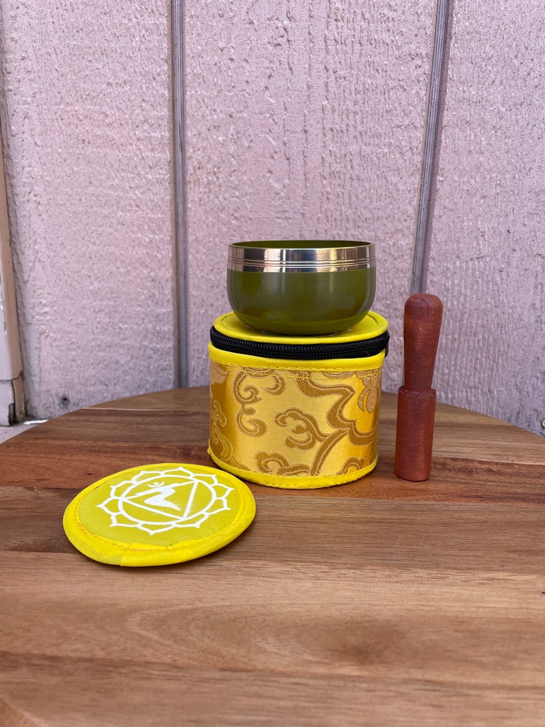 May include: A green singing bowl with a gold rim, a yellow fabric pouch with gold embroidery, and a wooden striker. The pouch has a white symbol on the top.