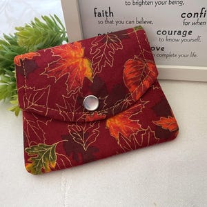 May include: A small fabric pouch featuring a deep red background with a pattern of autumn leaves in shades of orange, yellow, and brown. The pouch has a triangular flap secured by a single silver-toned snap button closure and visible stitching.