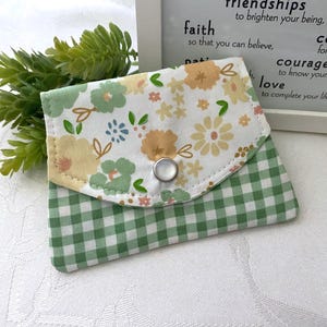 May include: A small, handmade wallet with a floral and gingham pattern. The top flap features a white background with yellow, green, and pink flowers. The bottom half is green and white gingham. A silver snap closure secures the wallet.