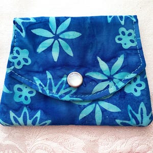 May include: A blue floral print wallet with a button closure. The wallet features a repeating pattern of turquoise flowers on a blue background. The edges are stitched with white thread.