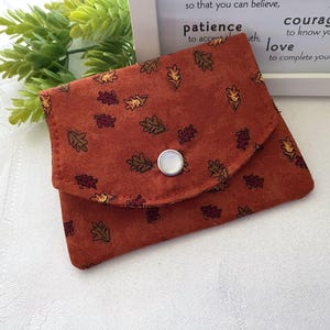 May include: A small, rectangular fabric pouch in a burnt orange color featuring a repeating pattern of small autumn leaves in shades of yellow, brown, and dark red. The pouch has a triangular flap secured by a single, centered white snap button.