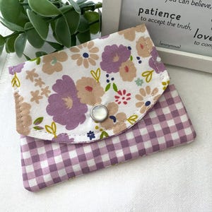 May include: A small, floral-patterned wallet with a gingham bottom. The top flap features a white background with purple, tan, and pink flowers. The bottom is a purple and white gingham pattern. A silver snap closure secures the wallet.