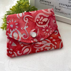 May include: A small, red batik fabric wallet with a white floral and swirl pattern. The wallet has a flap closure with a silver snap. The background includes a green plant and a framed inspirational quote.