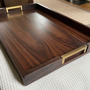 May include: A rectangular wooden serving tray with a rich, dark brown finish. The tray features brass-colored handles on each end. The wood grain is visible, adding to the tray's natural aesthetic. The tray is resting on a white textured surface.