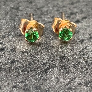 May include: Two gold stud earrings, each with a round, bright green gemstone. The earrings have a classic four-prong setting, highlighting the vivid green colour of the stones. The posts are visible, indicating a standard stud earring design.