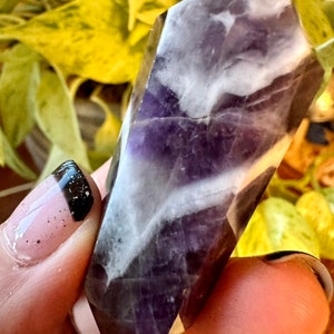 May include: A purple and white amethyst crystal point with a faceted shape. The crystal is smooth and polished.