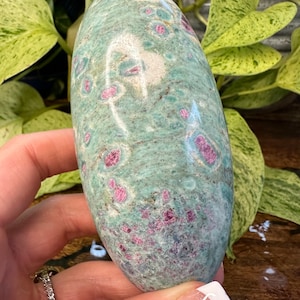 Peut inclure: Une pierre polie verte et rose de forme ovale lisse. La pierre présente un motif moucheté avec des inclusions roses et blanches.