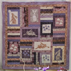 May include: A patchwork quilt with rabbit and floral motifs in shades of purple, brown, and cream. The quilt is displayed above a terracotta pot filled with vibrant pink and purple flowers. The quilt's dimensions are approximately 100 cm x 150 cm.