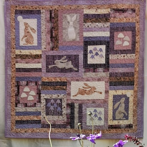 Pode incluir: Um cobertor de retalhos roxo e castanho com um padrão repetido de coelhos e cogumelos. O cobertor está pendurado numa parede branca com uma variedade de flores em vasos em primeiro plano.