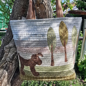 May include: A quilted tote bag featuring a brown dog and green trees design. The bag has brown straps and is made from a patchwork of different fabrics.