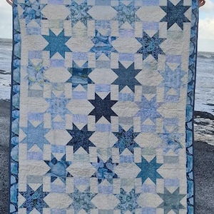 Puede incluir: Una colcha azul y blanca con un patrón de estrellas. La colcha está hecha de diferentes telas con una variedad de patrones azules y blancos. La colcha se exhibe en una playa.
