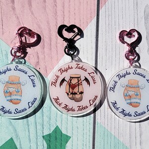 May include: Three round keychains with the text "Thick Thighs Saves Lives" and illustrations of thighs. One keychain has a pink heart-shaped clasp, another has a black heart-shaped clasp, and the third has a pink heart-shaped clasp.