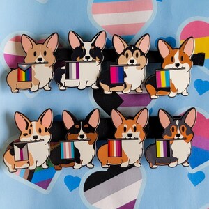 May include: A set of eight enamel pins shaped like Corgi dogs, each holding a different pride flag in its mouth. The pins feature various Corgi coat colors including tan, black, and white, with distinct pride flag patterns on each small rectangular banner.