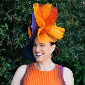 Könnte beinhalten: Eine Frau trägt ein leuchtend orangefarbenes und violettes Faltenkleid mit einem großen, farbenfrohen Hut. Der Hut ist aus gefaltetem Stoff in Orange-, Rot- und Violetttönen gefertigt.