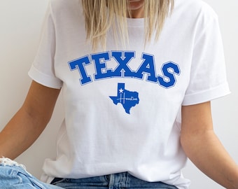 May include: White t-shirt with the word "TEXAS" in blue block letters across the chest. Below the word "TEXAS" is a blue outline of the state of Texas with a star and the word "Houston" inside.