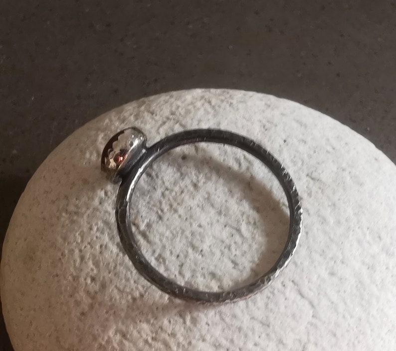 May include: A silver ring with a textured band and a round, silver bezel set with a small, red gemstone.