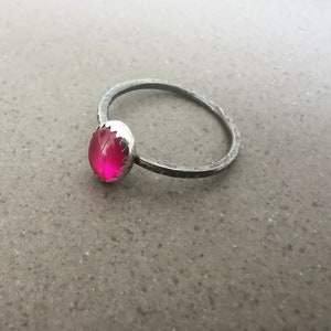 May include: A silver ring with a bright pink gemstone set in a scalloped bezel. The ring band is hammered and has a matte finish.