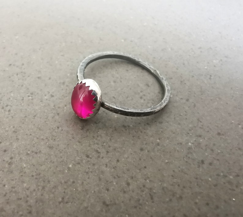 May include: A silver ring with a bright pink gemstone set in a scalloped bezel. The ring band is hammered and has a matte finish.