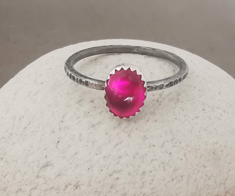 May include: A silver ring with a bright pink gemstone set in a textured silver bezel. The ring band is hammered and has a rustic look.