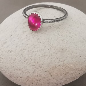 May include: A silver ring with a bright pink oval gemstone set in a silver bezel. The ring band is textured and has a hammered finish.