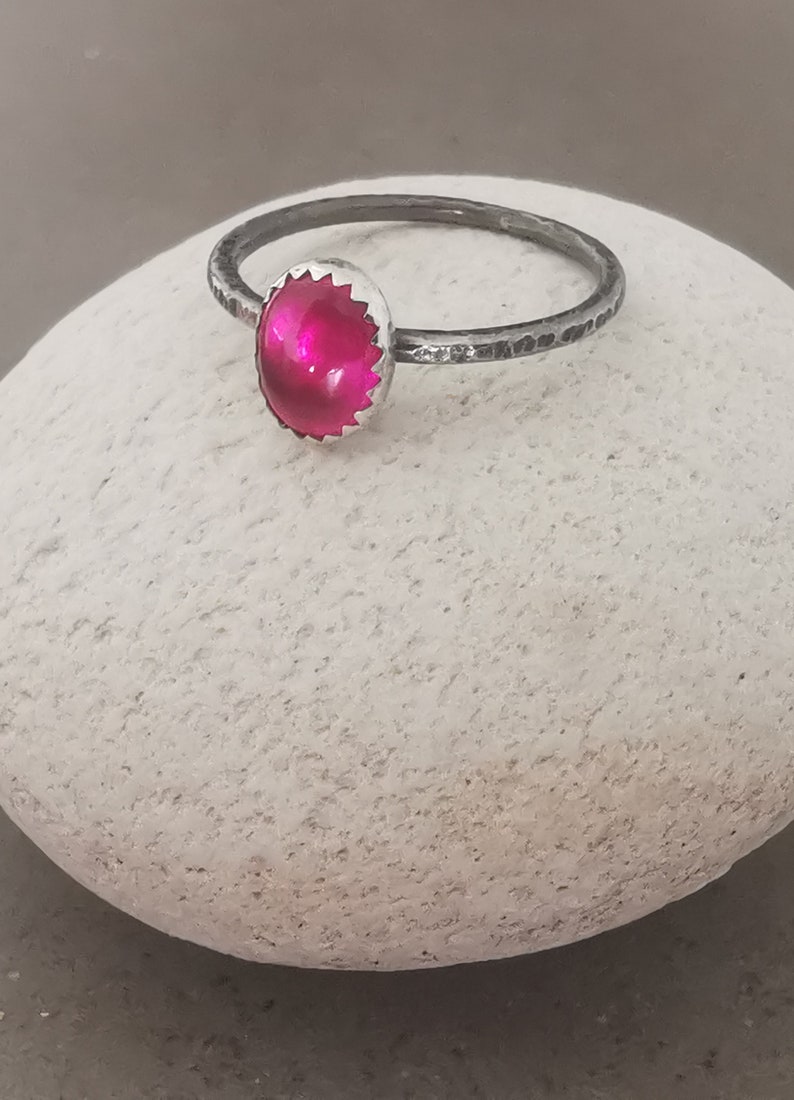 May include: A silver ring with a bright pink oval gemstone set in a silver bezel. The ring band is textured and has a hammered finish.