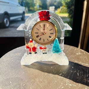 Op de afbeelding: Een boogvormige klok van heldere hars met een goudkleurige wijzerplaat en zwarte Romeinse cijfers. Binnenin de transparante structuur staan een miniatuurkerstman, een sneeuwpop en een blauwgroene kerstboom op een basis van witte glitters.
