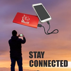 May include: A red power bank with a wolf logo and the word "WOLVES" printed on it. A white smartphone is plugged into the power bank with a black charging cable. The power bank and phone are floating in the air above a silhouette of a person standing on a rocky cliff. The sky is a vibrant orange and pink sunset. The text "STAY CONNECTED" is printed in black letters below the person.