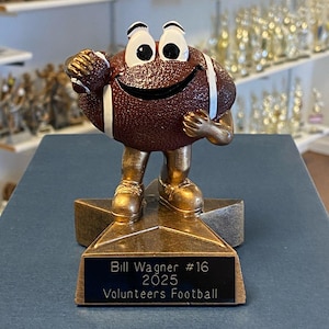 May include: A football-shaped trophy with a smiling face, arms, and legs, standing on a star-shaped base. The trophy is brown with white stripes and gold accents. The base has a plaque that reads "Bill Wagner #16 2025 Volunteers Football."